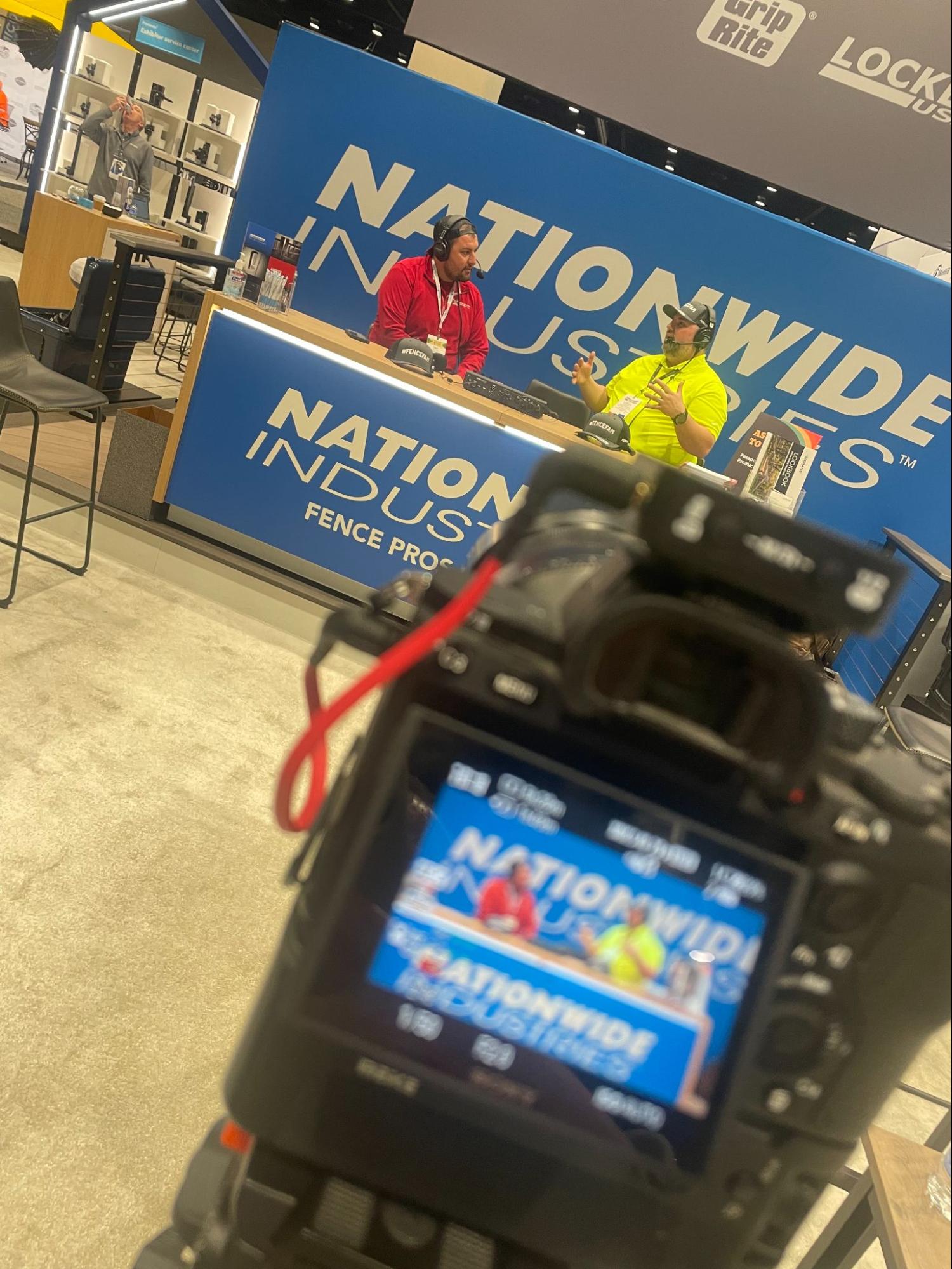 This picture shows a camera taking pictures of two people chatting at a Nationwide Industries booth.