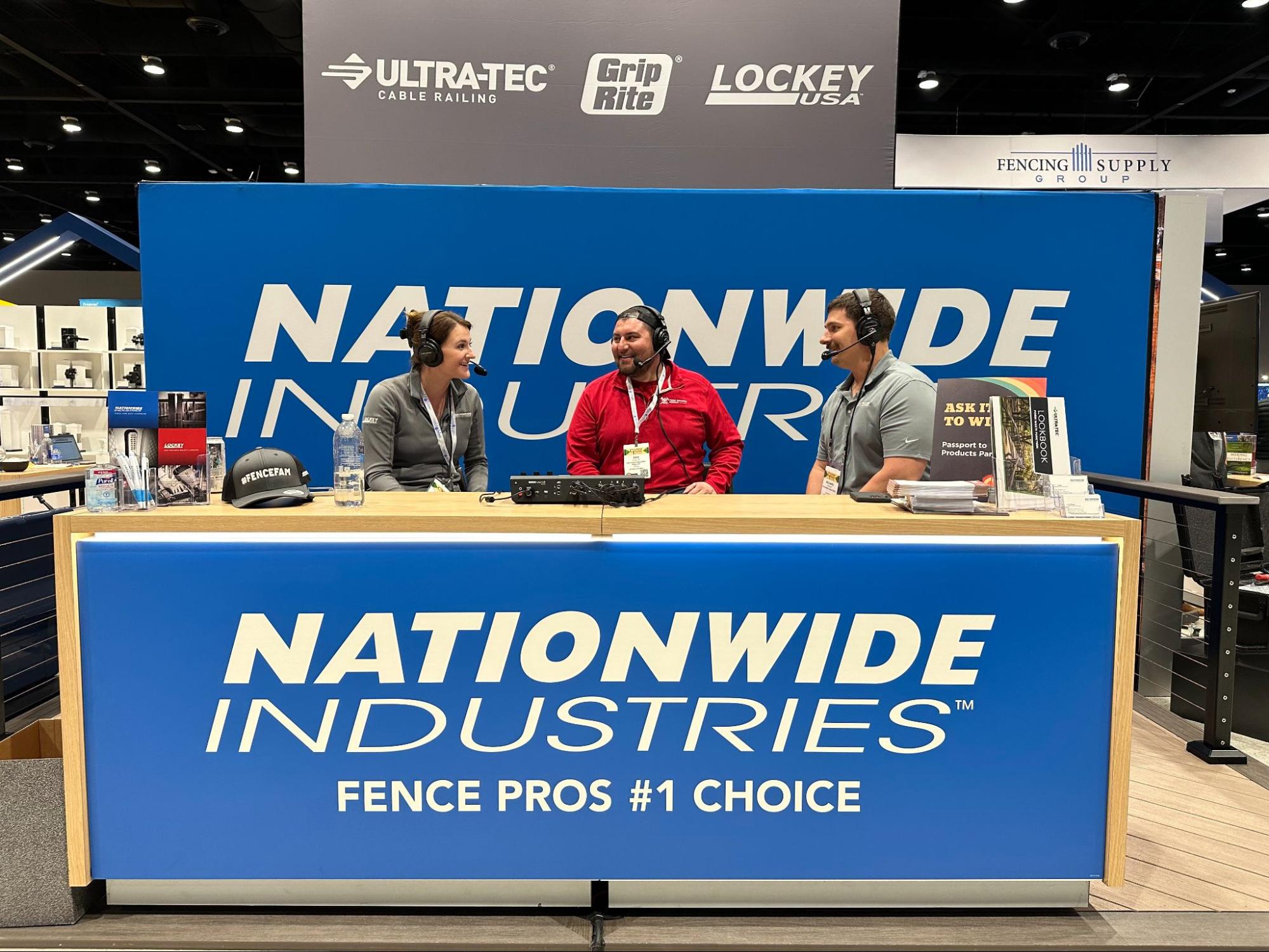 Three people are gathered at a Nationwide Industries exhibit behind a booth. They are talking to each other and smiling. 